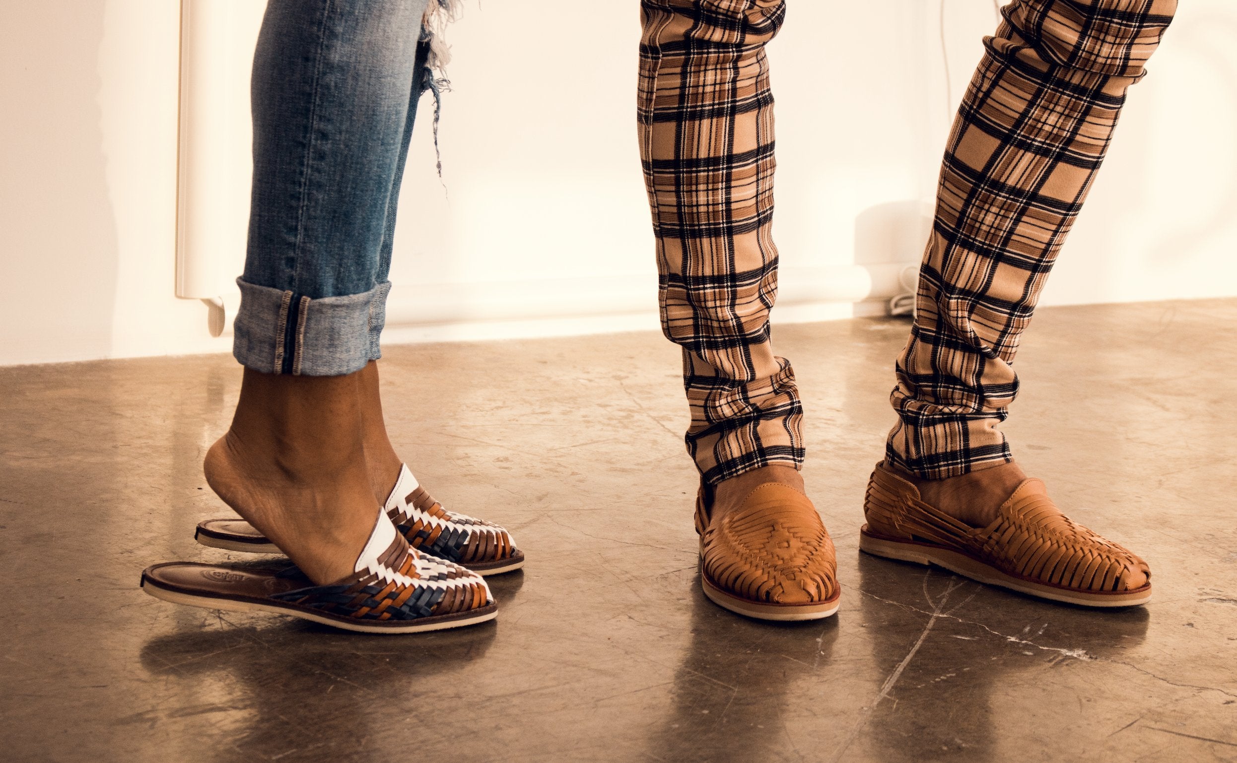 An image of two pairs of legs from the calf down, both wearing leather Mexican huaraches. One pair of legs is a woman, the other pair of legs is a man. 